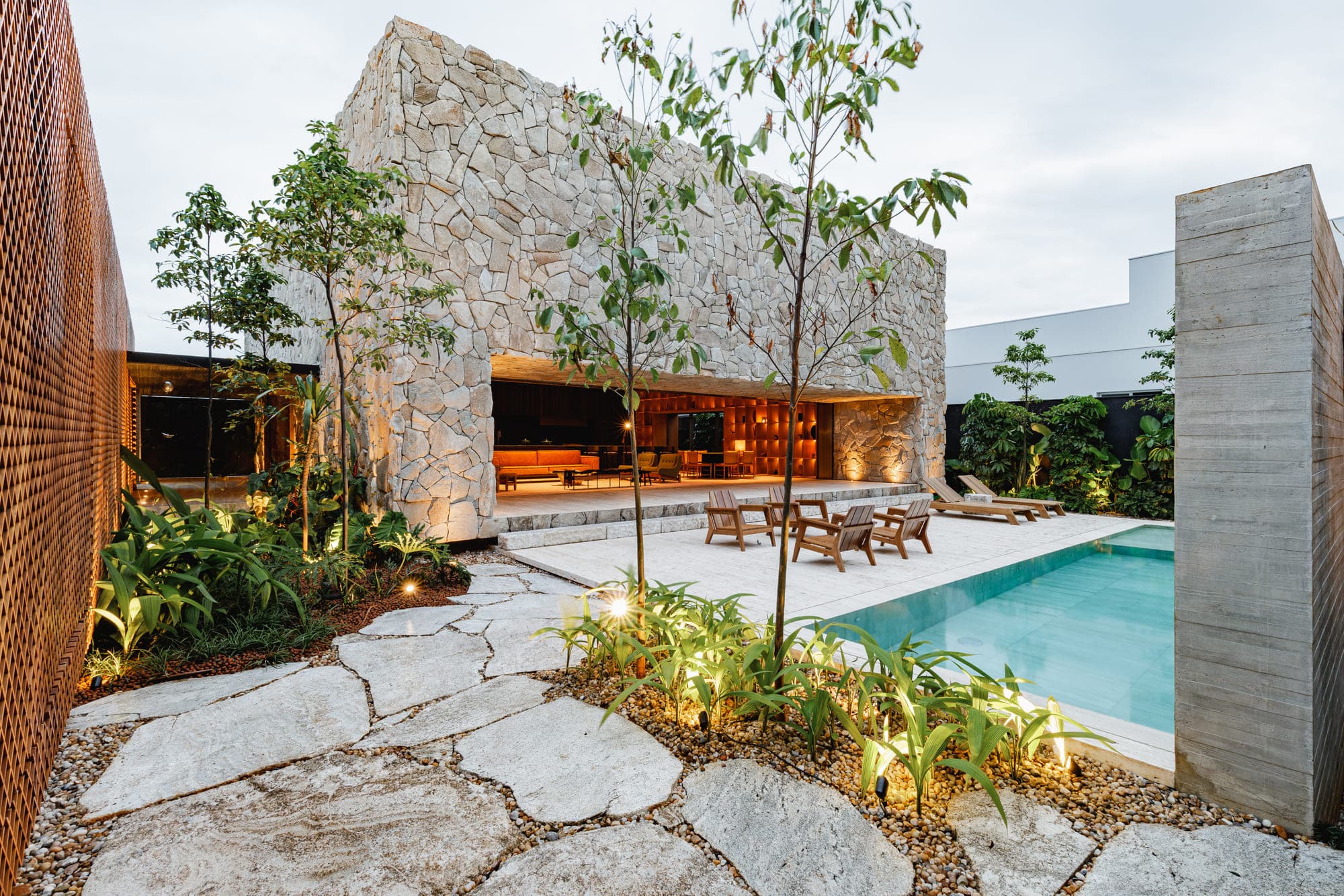 Pátio de casa com piscina, piso de pedra natural e árvores jovens junto ao muro de pedra