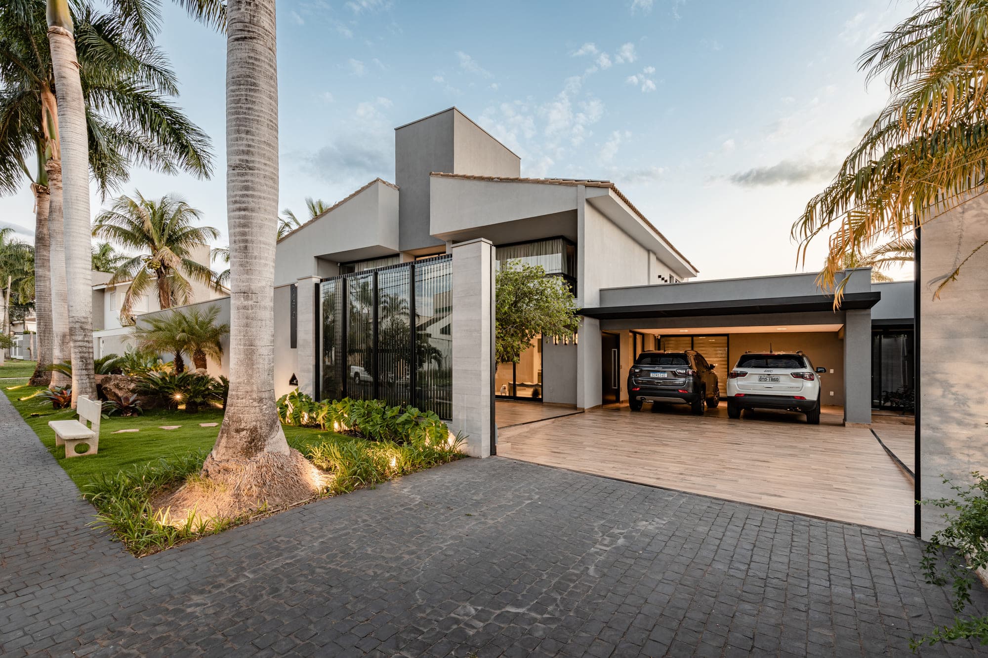 Casa branca ao anoitecer com garagem aberta, carros e palmeiras nas laterais