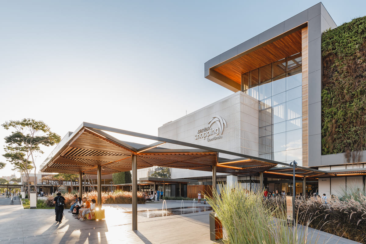 Entrada de shopping com cobertura em madeira, jardim e pessoas circulando
